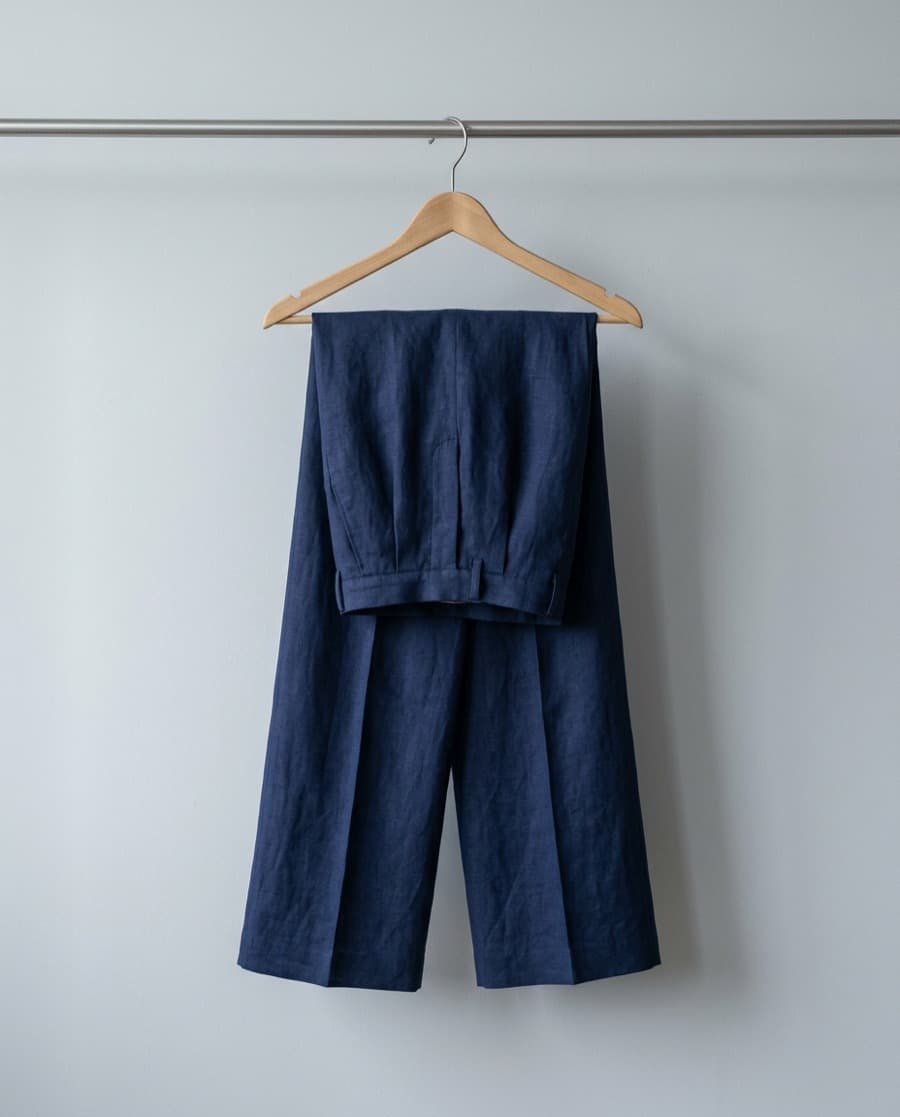 Navy wide-leg linen trousers folded over a wooden hanger against a pale grey wall