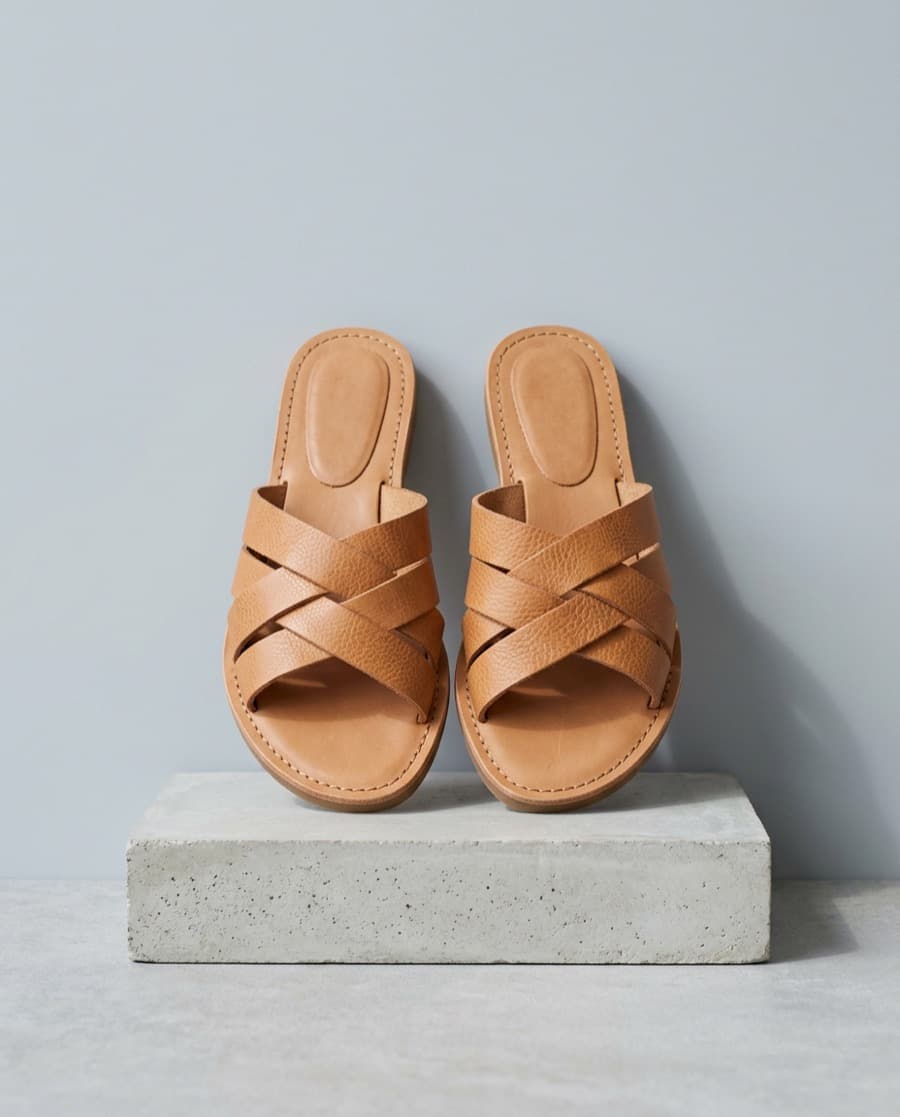 Pair of tan leather slides with crossed woven straps, standing on a concrete block