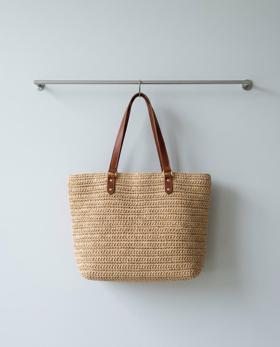 Natural woven raffia tote with brown leather handles, hanging from a steel rail